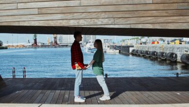 A couple holds hands on a wooden bridge overlooking a city waterfront, capturing a romantic moment.