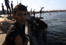 A boy sits by the water as children play, creating a lively scene in Gaza's coastal area.
