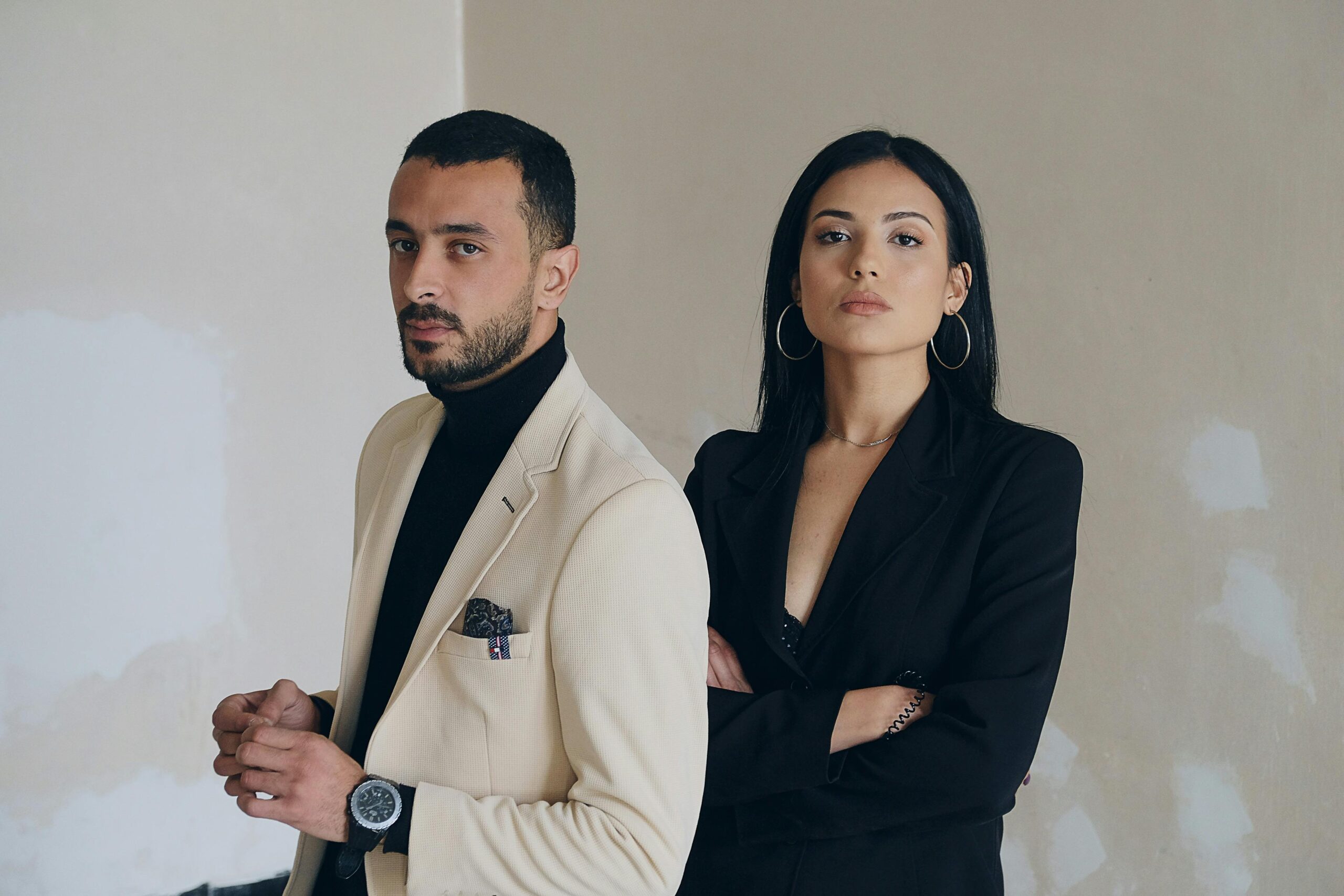 A man and woman posing confidently in stylish formal wear against a textured wall.